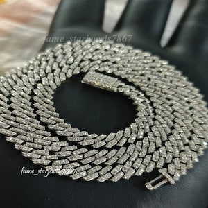 May include: A silver chain necklace with a diamond-like pattern. The chain is made up of small, rectangular links that are set close together. The chain is thick and heavy, and it has a clasp at the back. The chain is lying on a dark surface.