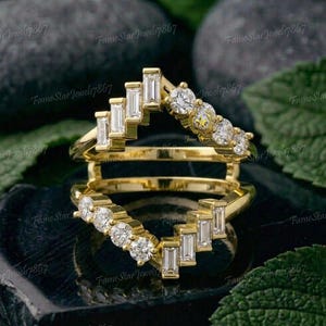 May include: A gold ring guard set featuring two chevron-shaped bands. Each band is adorned with a combination of clear baguette-cut and round brilliant-cut stones arranged in a stepped pattern along the top half of the polished gold metal bands.
