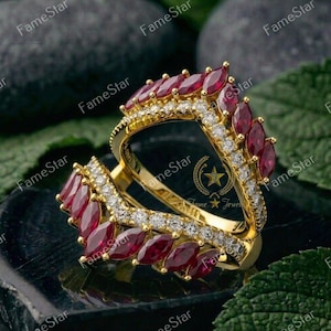 May include: A gold ring with a unique design, featuring marquise-cut red gemstones and small, sparkling white stones. The ring has a double-band design and is set against a dark background with green foliage.