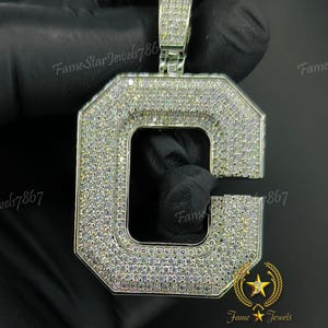 May include: A silver pendant in the shape of the letter C, completely covered in small, clear cubic zirconia stones. The pendant is on a black background.