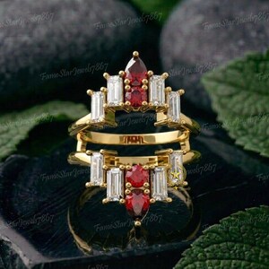 May include: A gold ring guard featuring two rows of stones. The top row has a pear-cut red stone, a round red stone, and four baguette-cut clear stones. The bottom row has a round red stone, a pear-cut red stone, and four baguette-cut clear stones.