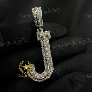 May include: A silver pendant in the shape of the letter J, encrusted with clear cubic zirconia stones. The pendant is held in a gloved hand.