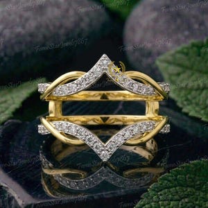 May include: A yellow gold ring guard featuring two chevron-shaped bands connected by a bridge. Each band displays a V-shaped design encrusted with small, clear, round-cut stones, creating a symmetrical, layered appearance with a polished finish.