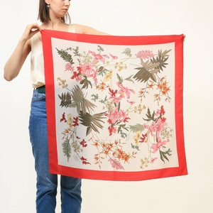 May include: A white silk scarf with a red border and a floral print of pink, yellow, and green flowers. The scarf is being held by a person.