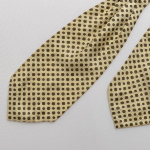 May include: A cream-coloured cravat with a repeating pattern of small brown squares. The cravat is made of a silky material and has a pointed end.