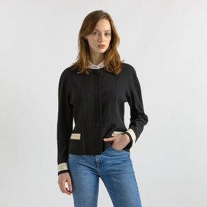 May include: A black cropped jacket with white trim on the cuffs and pockets. The jacket has a button closure and a collar. The model is wearing blue jeans.