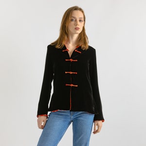 Puede incluir: Una chaqueta de terciopelo negro con ribetes rojos y cuello mandarín. La chaqueta tiene cinco botones en la parte delantera y mangas largas.