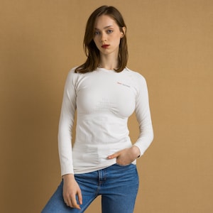 Puede incluir: Una camiseta blanca de manga larga con un diseño sin costuras y el texto "X-Bionic" en el pecho. La camiseta la lleva puesta una persona con el pelo largo y castaño y está de pie sobre un fondo marrón.