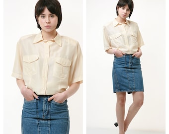 May include: A woman is wearing a light yellow button-down shirt with short sleeves and a blue denim skirt.
