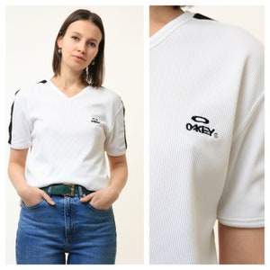 May include: White ribbed v-neck t-shirt with black and white striped detailing on the shoulders and a black "Oakley" logo on the front.