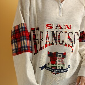 May include: A cream colored sweatshirt with a red and black plaid design on the sleeves. The front of the sweatshirt features an embroidered design of the Golden Gate Bridge with the text "SAN FRANCISCO" and "GOLDEN GATE BRIDGE".