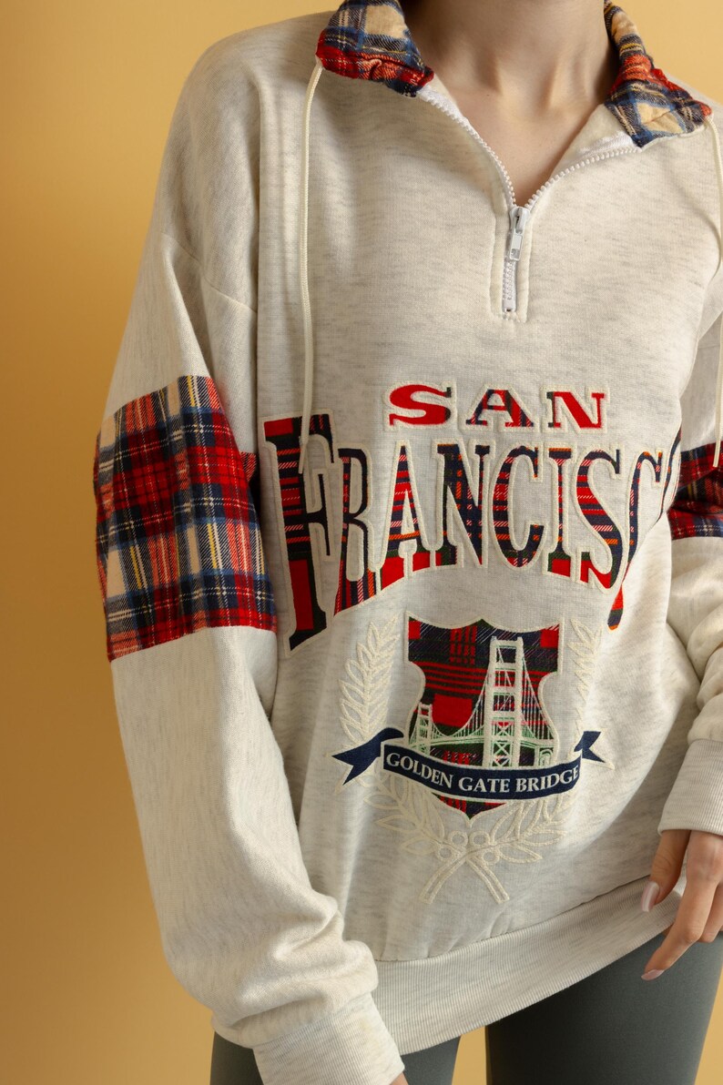 May include: A cream colored sweatshirt with a red and black plaid design on the sleeves. The front of the sweatshirt features an embroidered design of the Golden Gate Bridge with the text "SAN FRANCISCO" and "GOLDEN GATE BRIDGE".