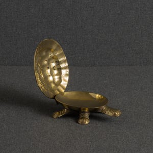 Könnte beinhalten: Eine polierte Messing-Schmuckschale in Form einer Schildkröte mit geöffnetem Panzer. Die Schildkröte hat eine glatte, goldfarbene Oberfläche, einen aufklappbaren Deckel, der aufrecht steht, und vier kleine Beine. Das Innere ist strukturiert.