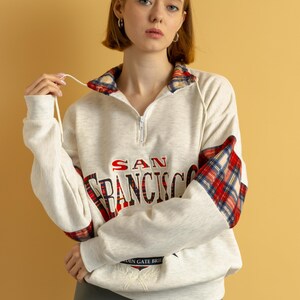 May include: A white sweatshirt with a red and black plaid trim on the sleeves and collar. The sweatshirt has the text "SAN FRANCISCO" printed on the front in red and blue lettering. The sweatshirt also has a small embroidered design of the Golden Gate Bridge on the front.