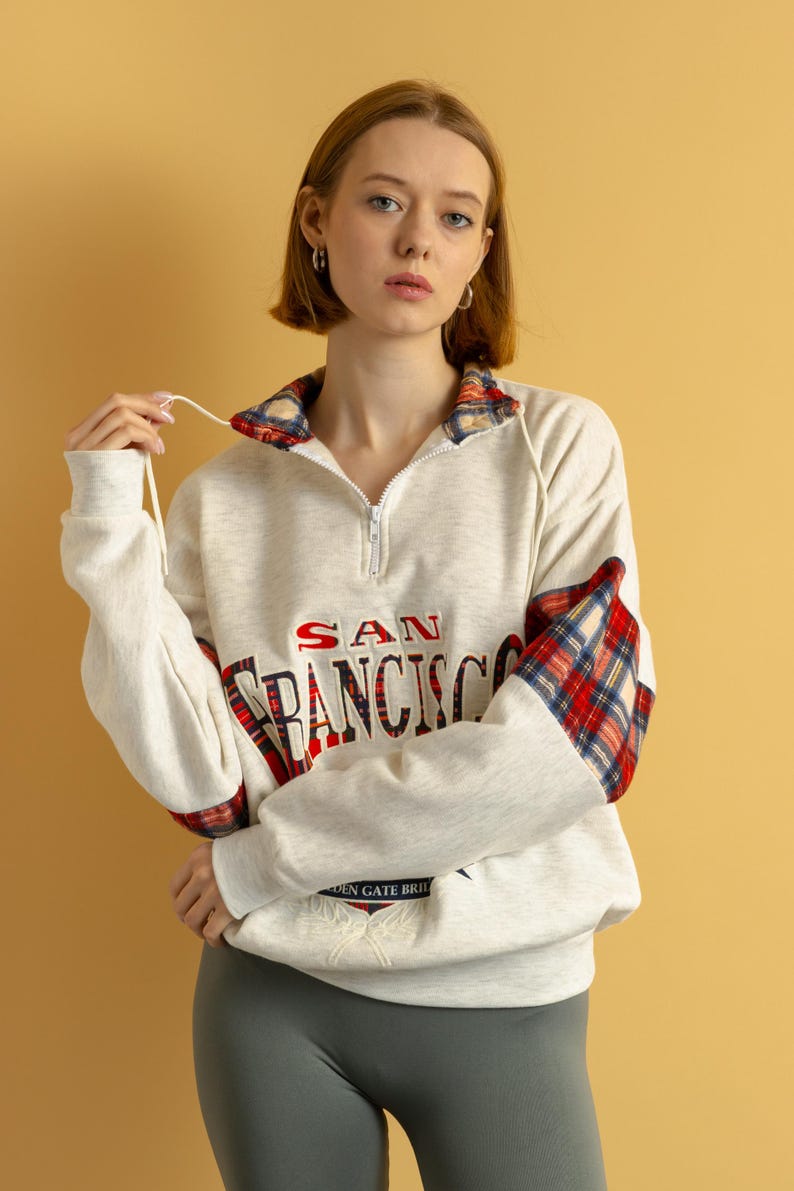 May include: A white sweatshirt with a red and black plaid trim on the sleeves and collar. The sweatshirt has the text "SAN FRANCISCO" printed on the front in red and blue lettering. The sweatshirt also has a small embroidered design of the Golden Gate Bridge on the front.