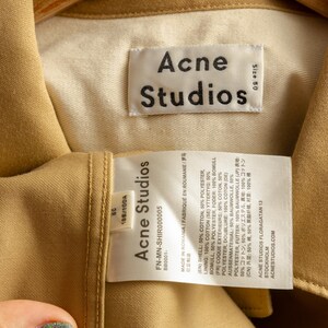 May include: A tan cotton jacket with a white label that reads "Acne Studios" and includes details about the fabric composition and size. The label also includes the text "FN-MN-SHIR000005" and "BB0001-".
