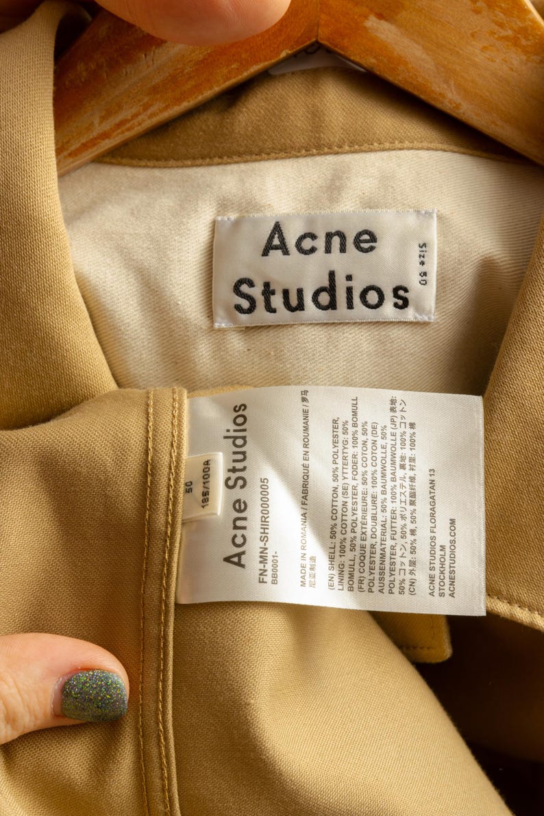 May include: A tan cotton jacket with a white label that reads "Acne Studios" and includes details about the fabric composition and size. The label also includes the text "FN-MN-SHIR000005" and "BB0001-".