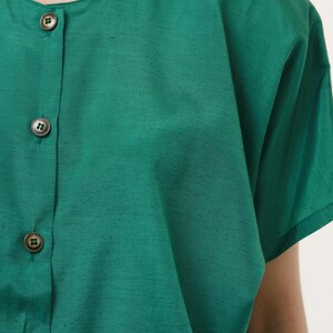 Puede incluir: Una camisa verde de botones con mangas cortas. La camisa tiene un cuello redondo y cuatro botones en la parte delantera.