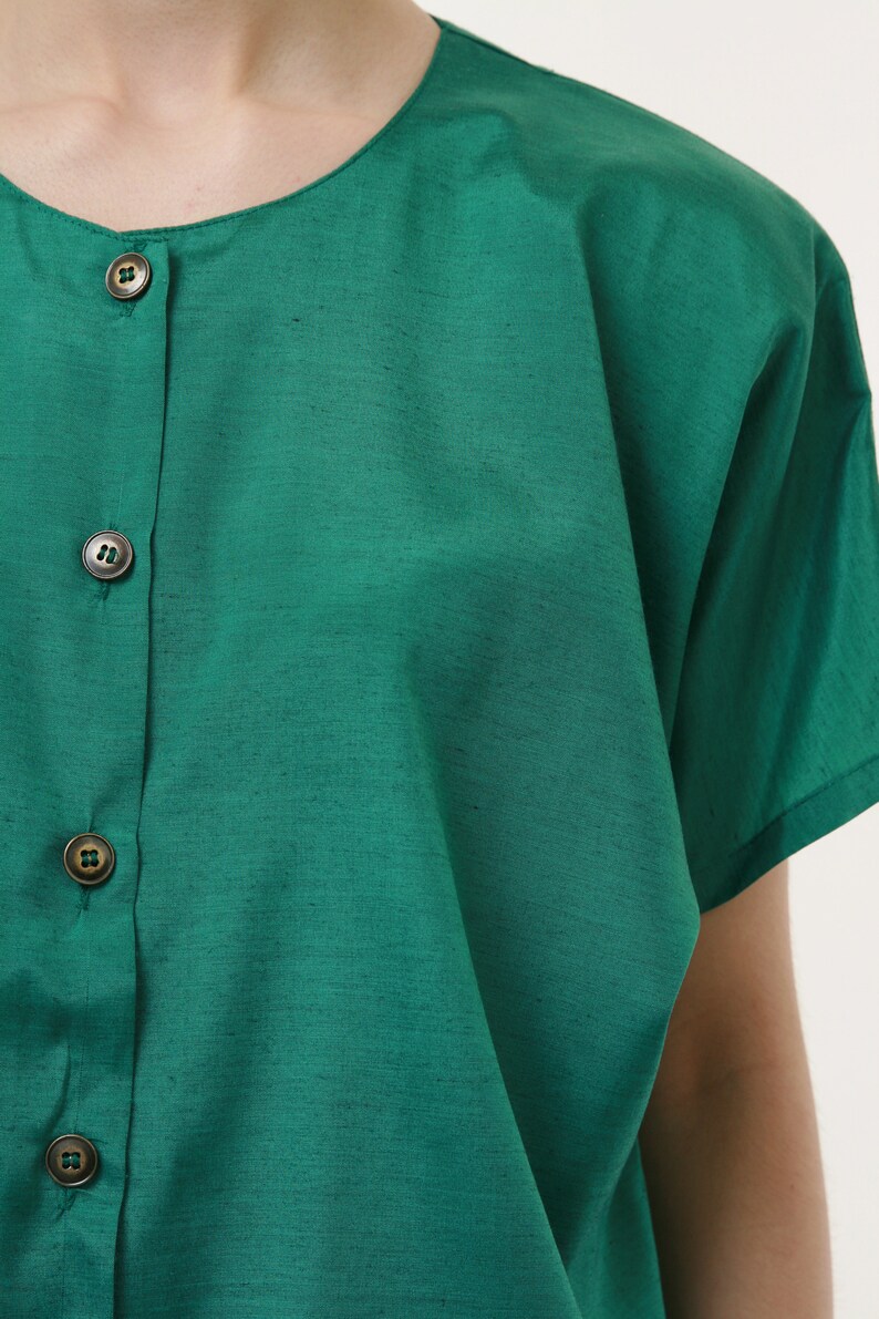 Puede incluir: Una camisa verde de botones con mangas cortas. La camisa tiene un cuello redondo y cuatro botones en la parte delantera.