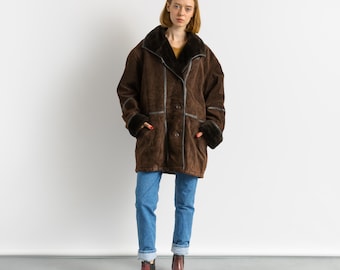 May include: A brown suede coat with a dark fur collar and cuffs. The coat has a button-up front and a relaxed fit. The person is wearing blue jeans and brown boots. The coat is a vintage style.