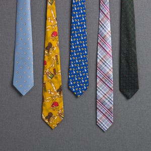 Puede incluir: Cinco corbatas estampadas de varios colores se muestran sobre un fondo gris. Las corbatas incluyen una corbata azul claro con lunares, una corbata amarilla con un patrón detallado, una corbata azul con un diseño repetido, una corbata a cuadros y una corbata verde estampada.
