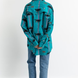 May include: A turquoise blue long-sleeved shirt with a colorful patterned design. The shirt has a button-down front and a relaxed fit.