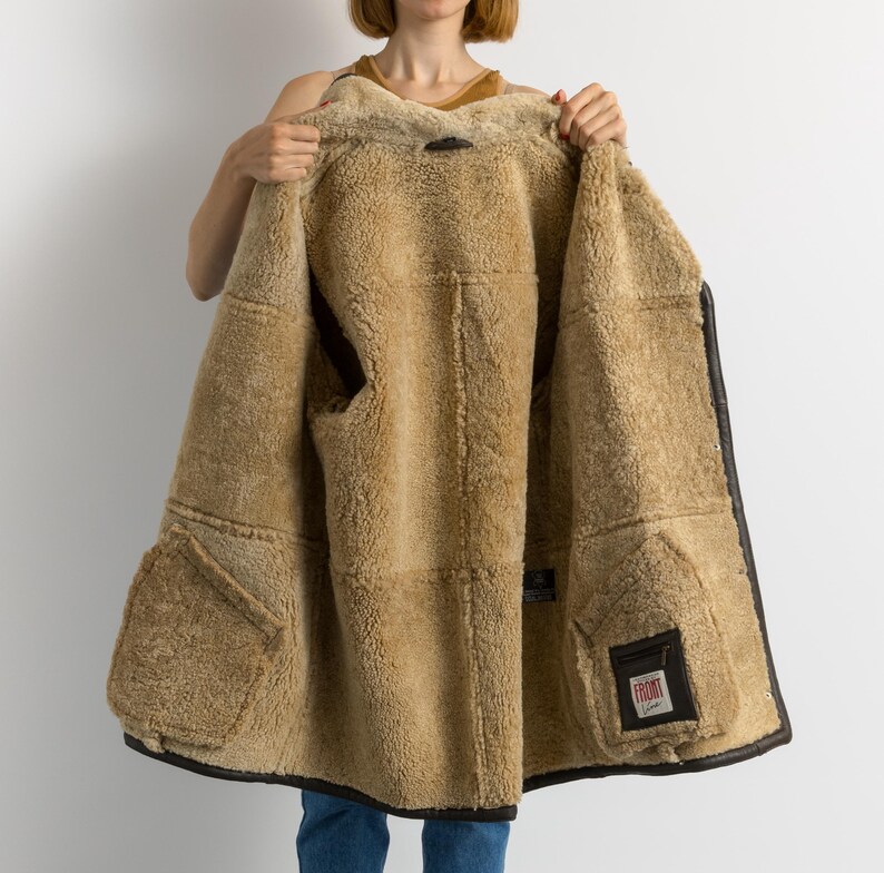May include: A brown shearling coat with a dark brown leather trim. The interior is a light tan color with two large pockets. The coat is open, revealing the soft, fluffy shearling lining. A small label is visible on the inside.