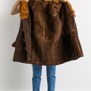 May include: A brown shearling coat with a fur collar is held open, revealing the plush interior. The coat features large buttons and a suede exterior. The model is wearing blue jeans and burgundy boots.
