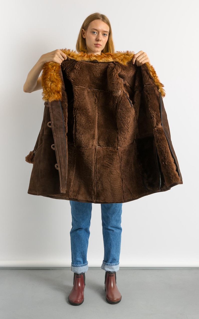 May include: A brown shearling coat with a fur collar is held open, revealing the plush interior. The coat features large buttons and a suede exterior. The model is wearing blue jeans and burgundy boots.