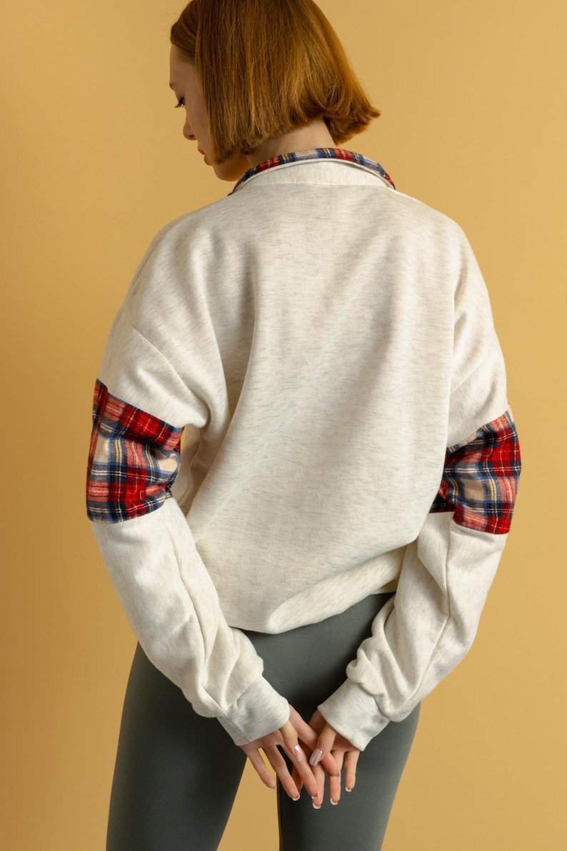 May include: A cream-colored sweatshirt with red and black plaid fabric on the sleeves. The sweatshirt has a relaxed fit and a crew neck.