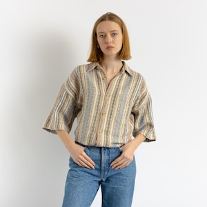 以下が含まれることがあります： ゆったりとしたシルエットのベージュとブルーのストライプ柄のボタンダウンシャツとブルーのジーンズを着用した女性