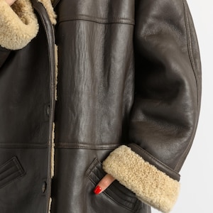 May include: A brown leather coat with a shearling lining and trim. The coat has a button closure, a notched collar, and pockets. The cuffs and collar are trimmed with beige shearling. The coat is a classic style.