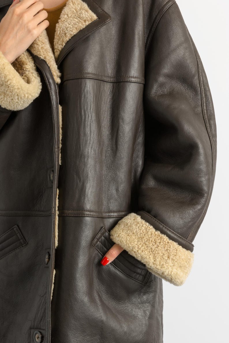 May include: A brown leather coat with a shearling lining and trim. The coat has a button closure, a notched collar, and pockets. The cuffs and collar are trimmed with beige shearling. The coat is a classic style.