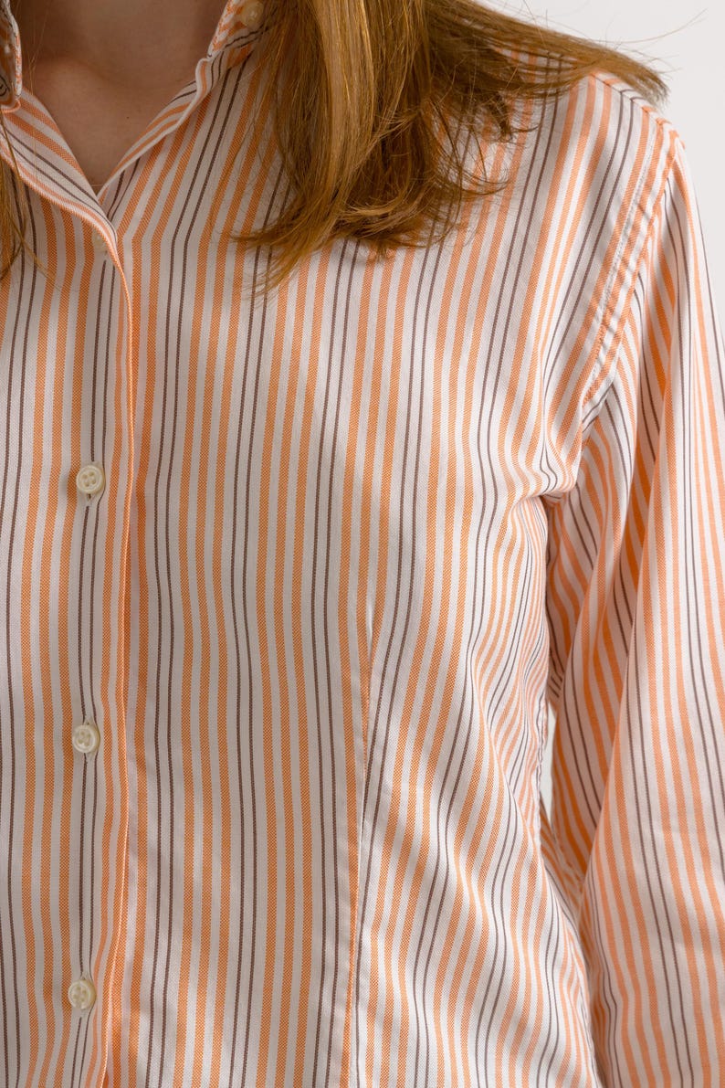 Puede incluir: Primer plano de una camisa de botones a rayas con rayas naranja y blancas. La camisa tiene un cuello cl&aacute;sico y mangas largas.