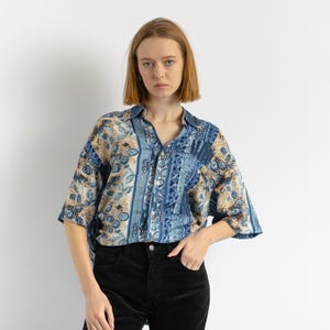 Puede incluir: Una mujer con una camisa de botones azul y marrón con manga larga. La camisa tiene un diseño de paisley. Lleva pantalones negros.