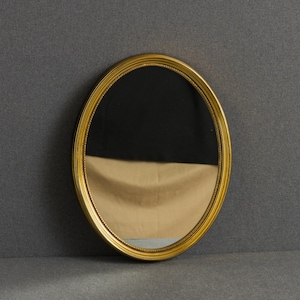 May include: An oval wall mirror featuring a polished gold-toned frame with a delicate beaded inner border. The reflective glass surface shows a clear view of the surrounding environment, capturing a reflection of a dark area and a light tan surface.