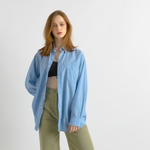 May include: A woman wearing a light blue oversized button-down shirt over a black tank top and olive green pants.