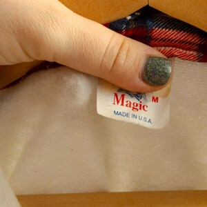 May include: A white tag with red text that reads "Magic" and "Made in U.S.A." with a size "M" label. The tag is attached to a red and blue plaid shirt.