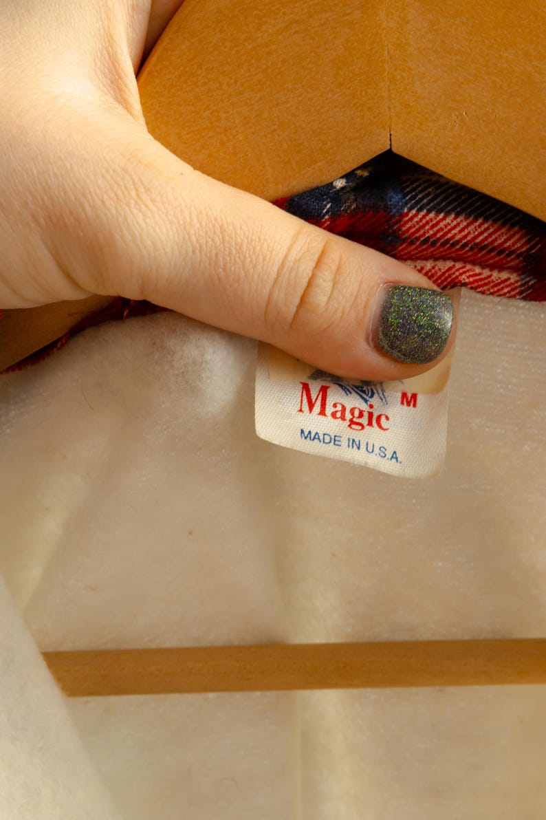 May include: A white tag with red text that reads "Magic" and "Made in U.S.A." with a size "M" label. The tag is attached to a red and blue plaid shirt.