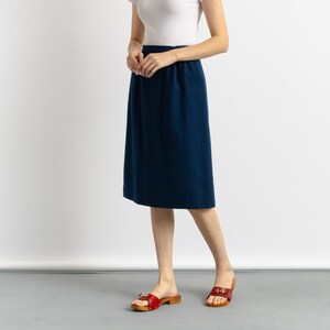 May include: A woman wearing a white t-shirt and a dark blue knee-length skirt. She is wearing red sandals with gold buckles.