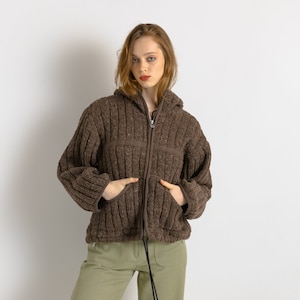 Könnte beinhalten: Eine braune Strickjacke mit Reißverschluss und Kapuze. Die Jacke hat eine gerippte Struktur und einen Reißverschluss. Das Model trägt die Jacke mit einer grünen Hose.
