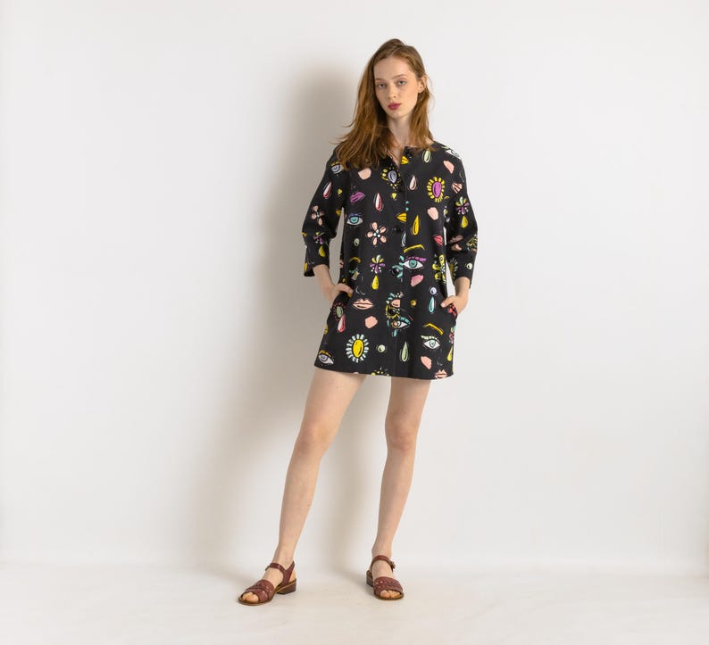 May include: A black button-down shirt dress with a colorful print of eyes, lips, and jewels. The dress has a relaxed fit and three-quarter sleeves.