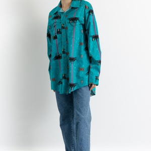 May include: A turquoise blue long-sleeved shirt with a colorful geometric pattern and black and white animal silhouettes. The shirt is worn with blue jeans and red sandals.
