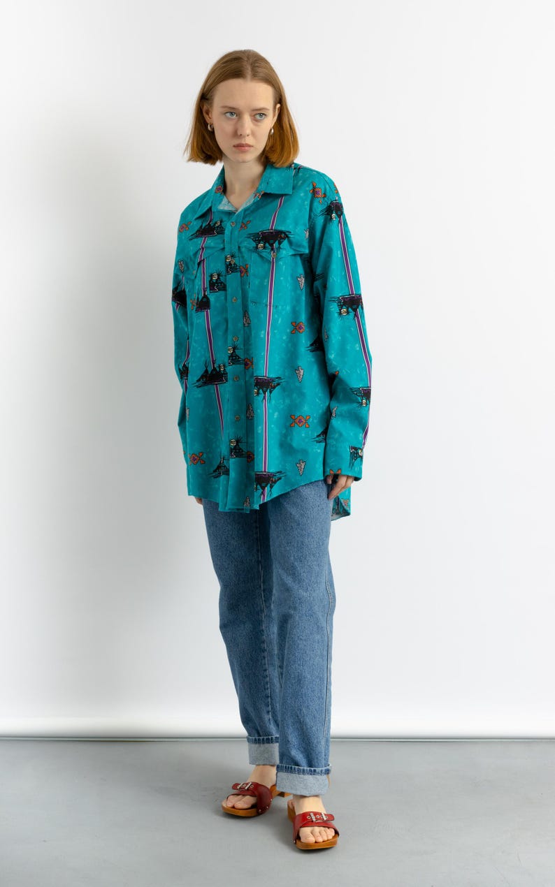 May include: A turquoise blue long-sleeved shirt with a colorful geometric pattern and black and white animal silhouettes. The shirt is worn with blue jeans and red sandals.