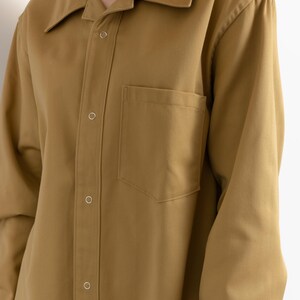 May include: A long-sleeved, collared shirt in a light brown color. The shirt has a button-up front and a single pocket on the left chest.