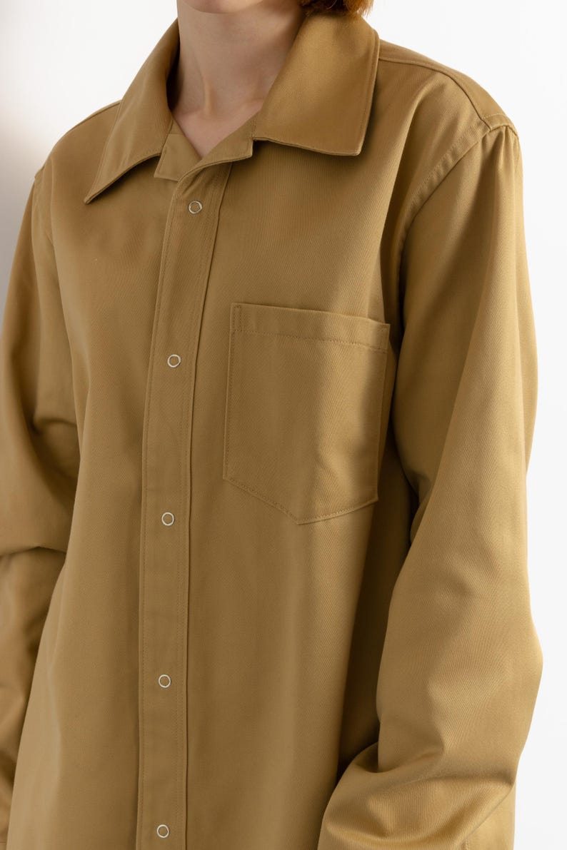 May include: A long-sleeved, collared shirt in a light brown color. The shirt has a button-up front and a single pocket on the left chest.