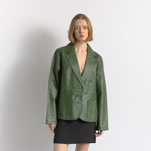 Op de afbeelding: Een groene leren blazer met een reverskraag, twee voorzakken en een sluiting met één knoop. De blazer wordt gecombineerd met een zwarte rok. Het model staat voor een witte achtergrond.