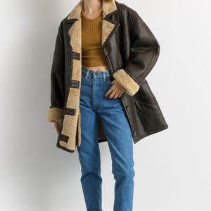 May include: A dark brown leather coat with a beige shearling lining and trim. The coat is worn over a mustard-colored top and blue jeans. The model is also wearing burgundy rain boots.