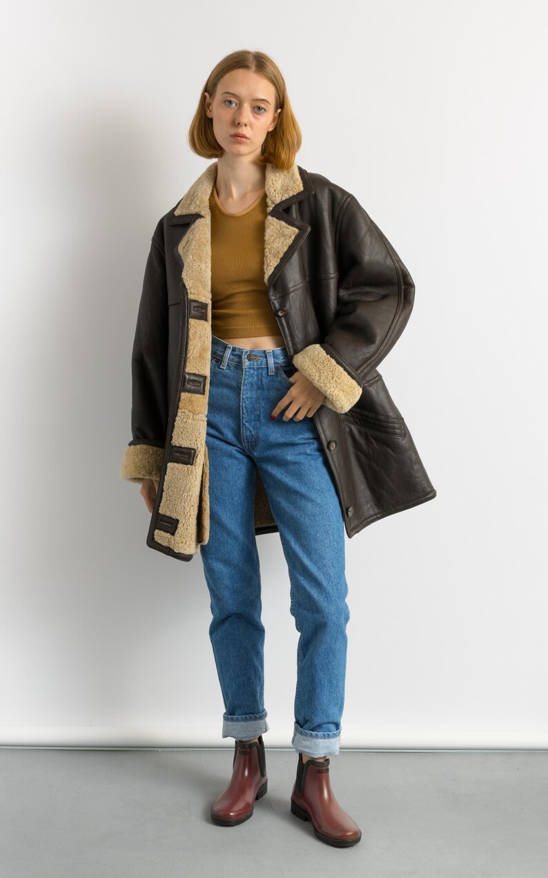 May include: A dark brown leather coat with a beige shearling lining and trim. The coat is worn over a mustard-colored top and blue jeans. The model is also wearing burgundy rain boots.