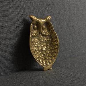 May include: A brass owl-shaped dish featuring a detailed, textured feather pattern across its surface. The metal tray has a distinct, rounded owl head at the top with two prominent circular eyes and a pointed beak, transitioning into a tapered body.